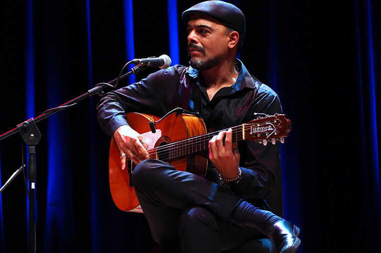 Ein Mann mit einer Schieberm&uuml;tze, der sitzt, Gitarre spielt und dabei in ein Mikrofon singt.