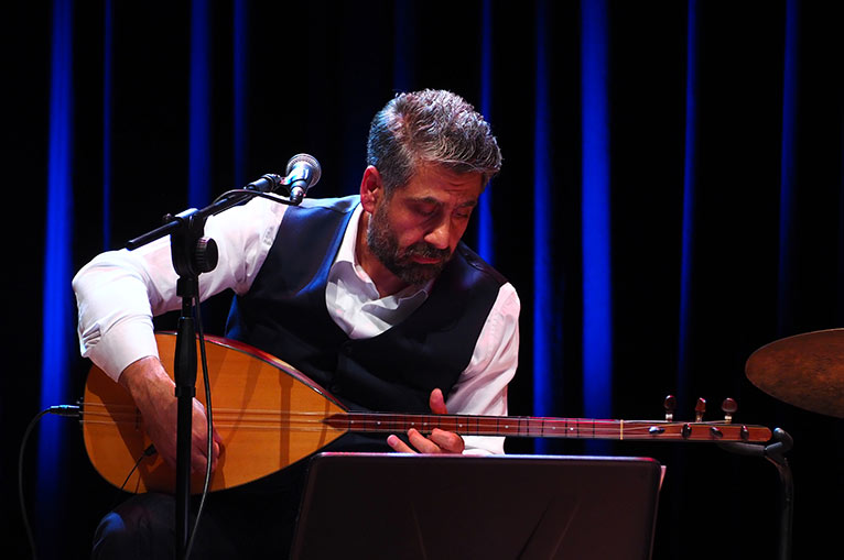 Ein Mann mit grauen Haaren, der ein gitarrenartiges Instrument spielt und dabei nach unten schaut. Vor ihm ein Mikrofon.