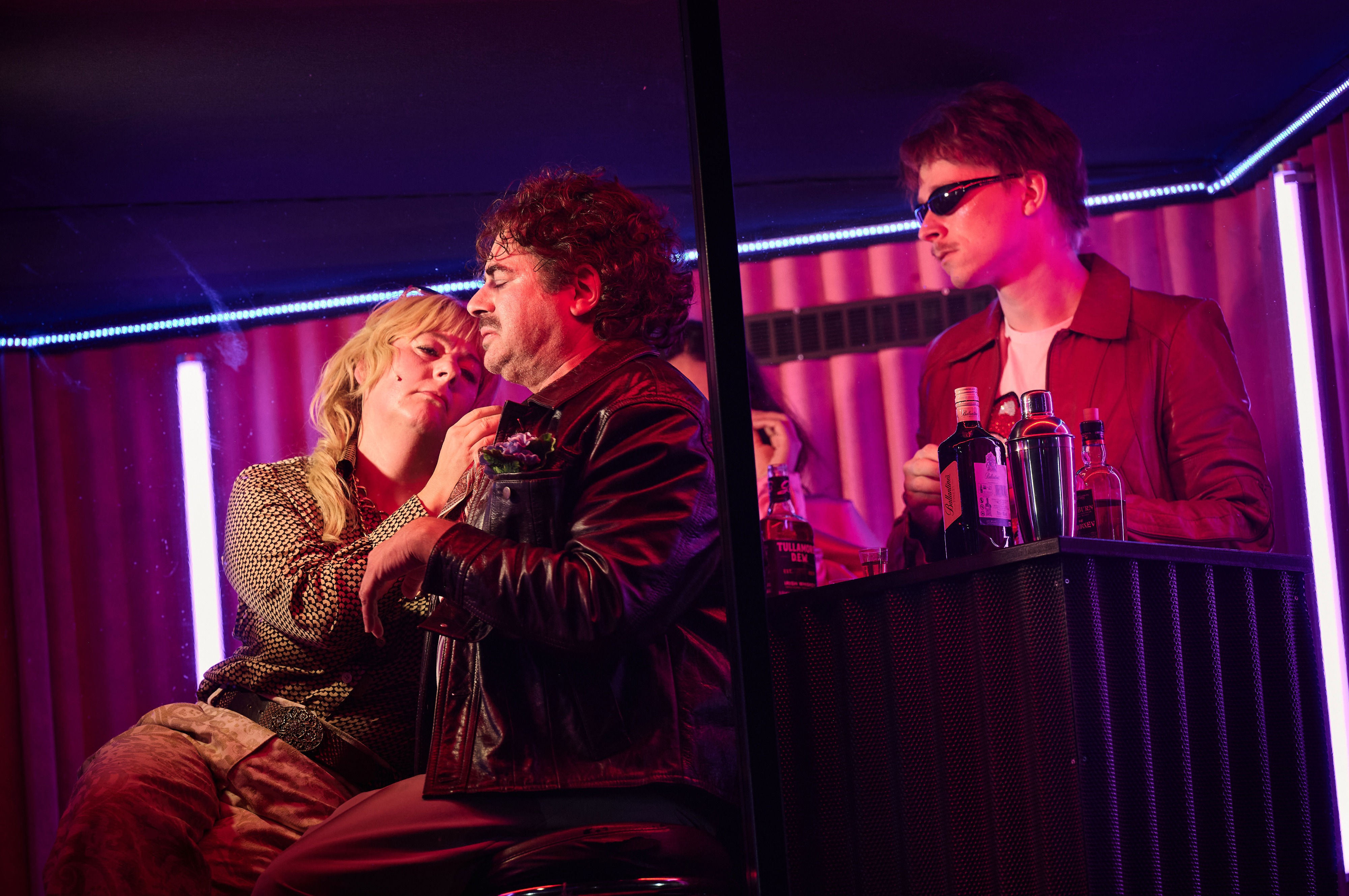 Ein Mann steht hinter einer Bar und tr&auml;gt eine Sonnenbrille. Davor sitzen zwei lehnt sich eine Schauspielerin an einen Schauspieler an.
